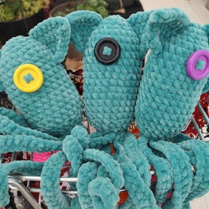 Puede incluir: Tres juguetes de pulpo de ganchillo turquesa con ojos de bot&oacute;n en amarillo, negro y morado. Los juguetes de peluche tienen largos tent&aacute;culos rizados y orejas grandes y ca&iacute;das. Se exhiben en un carrito de la compra de metal.