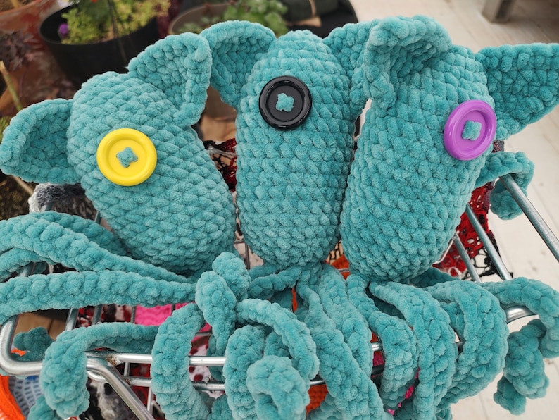 Puede incluir: Tres juguetes de pulpo de ganchillo turquesa con ojos de bot&oacute;n en amarillo, negro y morado. Los juguetes de peluche tienen largos tent&aacute;culos rizados y orejas grandes y ca&iacute;das. Se exhiben en un carrito de la compra de metal.