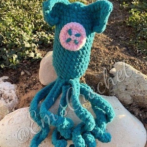 May include: A teal blue crocheted octopus with a pink button on its head. The octopus is sitting on a white rock. The text "PATTERN ONLY" is written below the octopus.