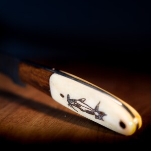 May include: A close-up of a wooden-handled knife with a bone-like handle. The handle has an intricate design of a fish etched into it.