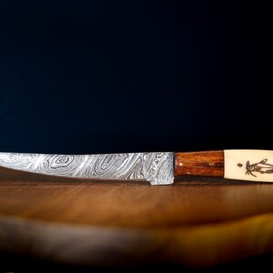 May include: A silver knife with a patterned blade and a wooden handle. The handle has a bone inlay with a fish design. The knife is resting on a wooden cutting board.