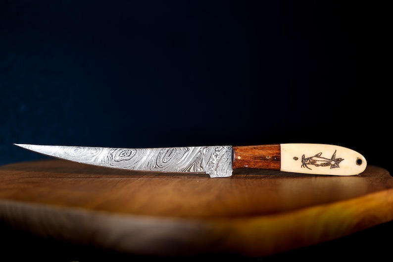May include: A silver knife with a patterned blade and a wooden handle. The handle has a bone inlay with a fish design. The knife is resting on a wooden cutting board.