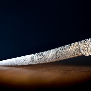 May include: A close-up of a silver knife with a patterned blade and a wooden handle. The knife is lying on a wooden surface.