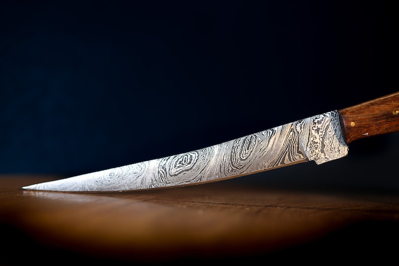 May include: A close-up of a silver knife with a patterned blade and a wooden handle. The knife is lying on a wooden surface.