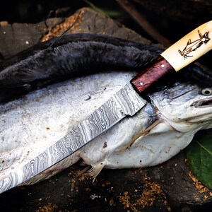 May include: A Damascus steel knife with a wooden handle and a bone handle with a carved catfish design. The knife is resting on top of a catfish.