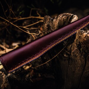May include: A brown leather knife sheath with a decorative stitching pattern and a white handle. The sheath is lying on a piece of weathered wood.