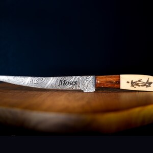 May include: A silver knife with a patterned blade and a wooden handle. The handle is made of two pieces of wood, one dark brown and one light brown. The knife has a bone-like tip with a carved design of a fish. The knife is engraved with the word "Moses".