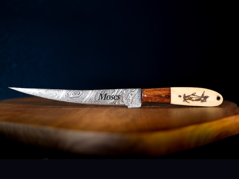 May include: A silver knife with a patterned blade and a wooden handle. The handle is made of two pieces of wood, one dark brown and one light brown. The knife has a bone-like tip with a carved design of a fish. The knife is engraved with the word "Moses".