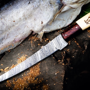 May include: A long, curved, stainless steel knife with a damascus pattern blade and a wooden handle. The handle has a bone inlay with a fish design. The knife is resting on a wooden surface with a fish skin beside it.