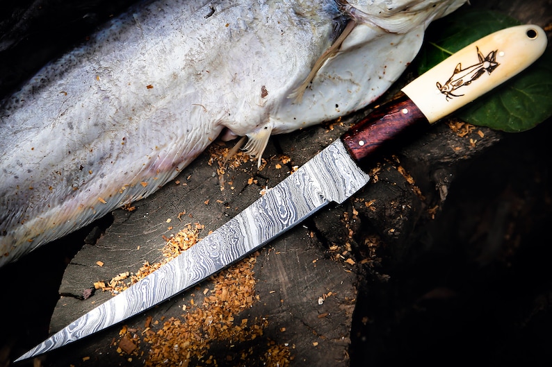 May include: A long, curved, stainless steel knife with a damascus pattern blade and a wooden handle. The handle has a bone inlay with a fish design. The knife is resting on a wooden surface with a fish skin beside it.