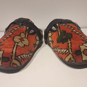 Könnte beinhalten: Ein Paar rote, schwarze und cremefarbene gemusterte Clogs mit einem schwarzen Lederriemen über dem Oberteil.