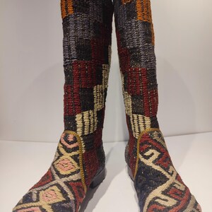 Könnte beinhalten: Ein Paar kniehohe Stiefel mit einem farbenfrohen, geometrischen Muster. Die Stiefel sind aus einem gewebten Stoff gefertigt und haben eine schwarze Gummisohle.