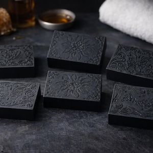 May include: Six dark gray rectangular soaps with ornate floral and scroll designs are arranged on a dark surface. A honeycomb, a small bowl, and a white towel are in the background. The soaps have detailed embossed patterns.