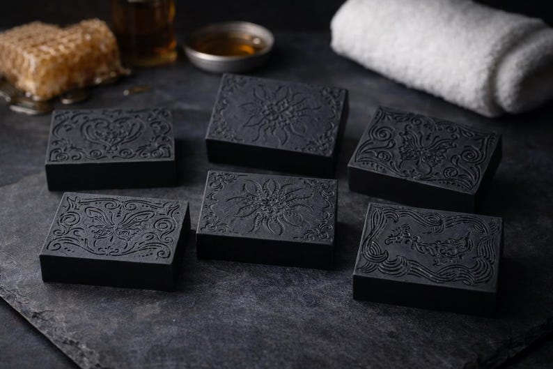 May include: Six dark gray rectangular soaps with ornate floral and scroll designs are arranged on a dark surface. A honeycomb, a small bowl, and a white towel are in the background. The soaps have detailed embossed patterns.