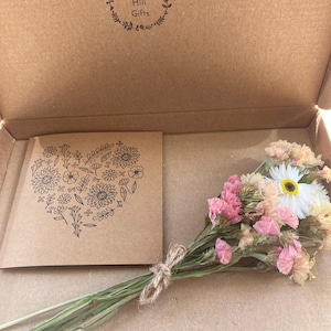 May include: A small bouquet of dried flowers in pink, white, and yellow, tied with twine, sits inside a cardboard box. A card with a floral heart design is also in the box. The box is stamped with the text "Wildflower Hill Gifts".