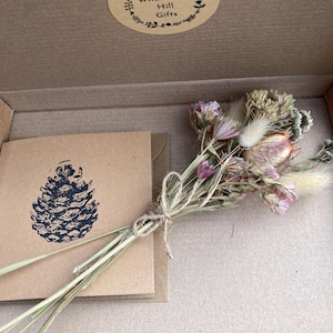 May include: A small bouquet of dried flowers in shades of pink, white, and brown, tied with twine. The flowers are in a cardboard box with a card that says "Wildflower Hill Gifts" and a pine cone illustration.