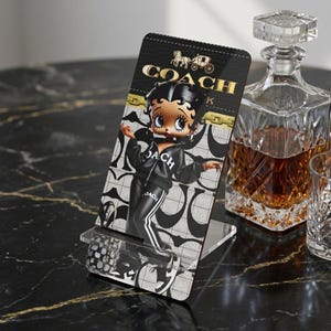 Può includere: Un supporto rettangolare in acrilico per telefono con una grafica di Betty Boop che indossa una tuta nera con il logo Coach. Lo sfondo presenta un motivo ripetuto in bianco e nero con dettagli a catena dorati e il nome Coach in oro.