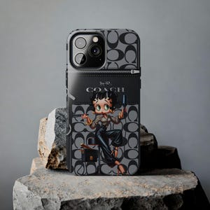 May include: A black and grey phone case featuring a cartoon image of Betty Boop holding a hairdryer. The case has a Coach logo and a black and grey pattern.