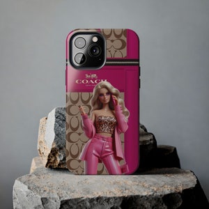 May include: A pink and brown iPhone case with a zipper detail and a graphic of a blonde doll wearing a pink outfit and holding a phone. The case has the Coach logo and the words "New York" printed on it.