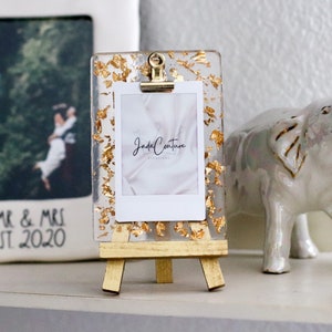 May include: A small gold picture frame with a white photo inside. The frame is on a wooden easel and has gold leaf accents. The photo is of a couple in front of a waterfall. The text "Jade Couture Photography" is visible on the photo.