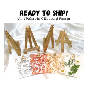 May include: Mini Polaroid clipboard frames with gold-painted wooden easels. The clipboards are clear with decorative elements, including silver and gold leaf, and dried flowers. The text "READY TO SHIP!" and "Mini Polaroid Clipboard Frames" are visible.