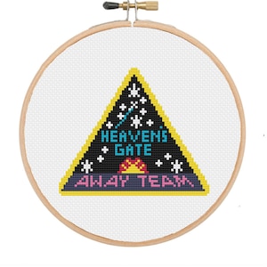 May include: A cross-stitch embroidery hoop featuring a triangular design with the words "HEAVENS GATE AWAY TEAM". The design includes stars, a sun, and a yellow border. The brand name "BlancheStitches" is above the hoop.