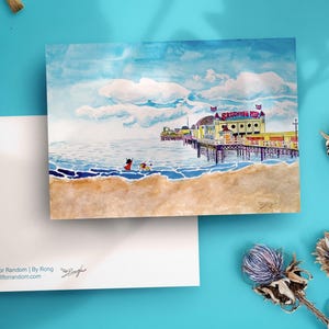 Könnte beinhalten: Aquarell-Kunstkarte mit Brighton Pier. Die Karte zeigt eine Strandszene mit Pier, Meer und Himmel. Die Karte wird mit einer leeren Karte und Pinseln auf einer blauen Oberfläche kombiniert.