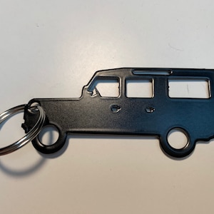 Könnte beinhalten: Schwarzer Metall-Schlüsselanhänger in Form eines viertürigen SUV. Der Schlüsselanhänger hat einen silbernen Schlüsselring. Das SUV-Design umfasst Details wie Fenster, Räder und einen Dachgepäckträger. Der Schlüsselanhänger ist ein solides, ausgeschnittenes Design.