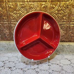 May include: A red ceramic divided serving dish with a white rim. The dish is divided into three sections and is sitting on a white crocheted doily.