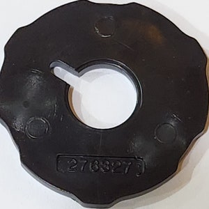 May include: A black sewing machine bobbin case with a central opening and a notched outer edge. The number "276327" is printed on the surface. The case is made of a durable material and is designed for sewing machine use.