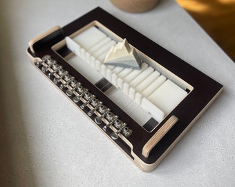 May include: A dark brown wooden soap cutting board with a white plastic insert and a row of metal knobs. A triangular piece of soap is resting on the board.