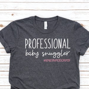Peut inclure: Un t-shirt gris fonc&eacute; avec le texte "PROFESSIONAL baby snuggler #NEWBORNPHOTOGRAPHER" en lettres blanches. Le hashtag est en rose.