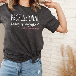 Peut inclure: Un t-shirt gris fonc&eacute; avec le texte "PROFESSIONAL baby snuggler #NEWBORNPHOTOGRAPHER" en blanc. Le texte "snuggler" est en rose.