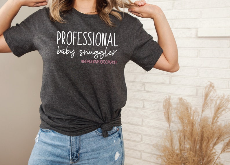 Peut inclure: Un t-shirt gris fonc&eacute; avec le texte "PROFESSIONAL baby snuggler #NEWBORNPHOTOGRAPHER" en blanc. Le texte "snuggler" est en rose.