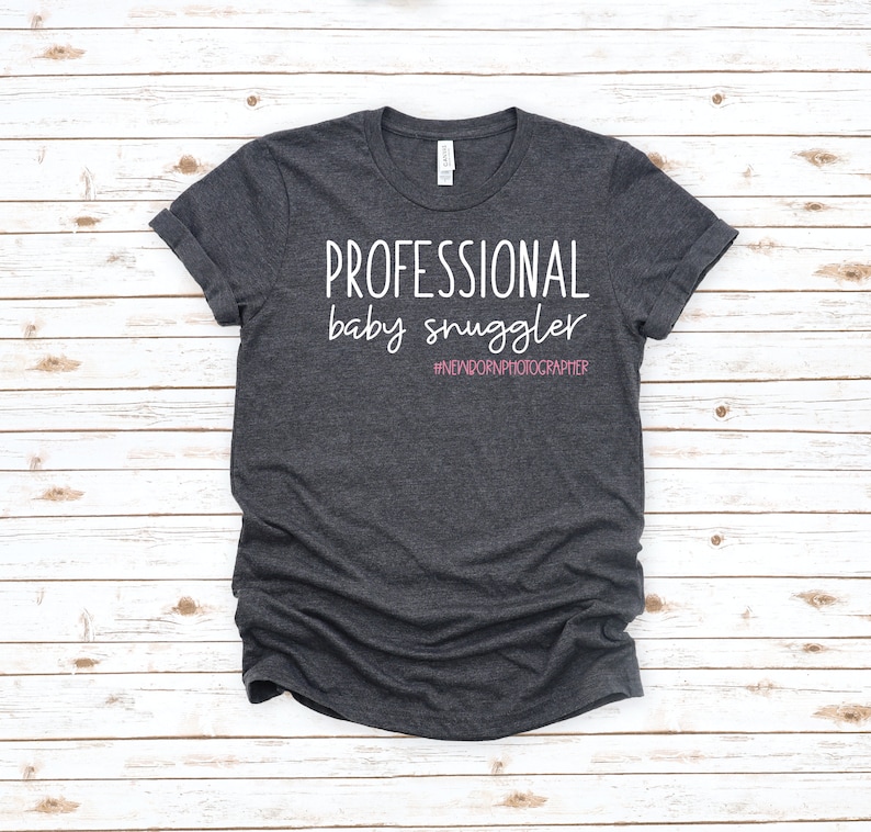 Peut inclure: Un t-shirt gris fonc&eacute; avec le texte "PROFESSIONAL baby snuggler #NEWBORNPHOTOGRAPHER" en lettres blanches. Le hashtag est en rose.