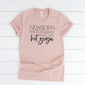 Peut inclure: Un t-shirt rose p&acirc;le avec le texte "NEWBORN PHOTOGRAPHY IS MY HOT YOGA" imprim&eacute; en noir.