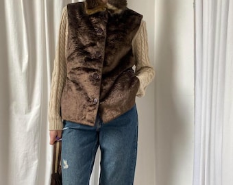 May include: A brown faux fur vest with a brown knit collar. The vest has a button closure and is worn over a white knit sweater.