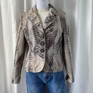 May include: A tan blazer with a unique animal print pattern featuring stripes and spots. The blazer has three decorative buttons, two front pockets, and a ruffled collar. The garment is paired with blue jeans.