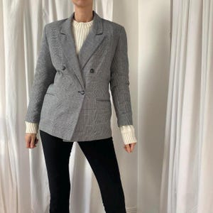 Può includere: Un blazer a quadri grigio con un design doppiopetto. Il blazer ha un revers a lancia, due tasche anteriori e una chiusura con un solo bottone. La modella indossa un maglione bianco e pantaloni neri.