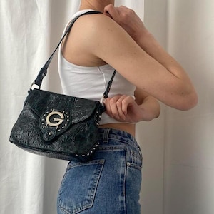 May include: A black leather shoulder bag with a silver buckle and a textured design. The bag is worn by a person wearing a white tank top and blue jeans.