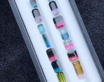 May include: A selection of rectangular gemstones presented in a black box with a white lining. The gemstones exhibit a range of colours, including blue, pink, green, and yellow. The stones are arranged in rows, highlighting their diverse hues and cuts.