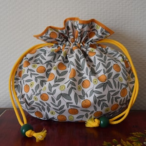 Puede incluir: Una bolsa de cordón blanco con un estampado floral amarillo y naranja con naranjas y hojas. La bolsa tiene un cordón de ajuste amarillo y dos borlas amarillas.
