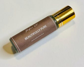 May include: A cylindrical glass perfume oil roller bottle with a metallic gold cap. The label is brown with white text that reads Perfume Oil and BEAUTIFULLY PURE, featuring a thin white horizontal line below the text on the bottle.