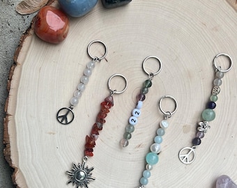 Puede incluir: Cinco llaveros de cuentas con diferentes dijes, incluyendo un signo de paz, un sol, una estrella de mar y una flor. Los llaveros se muestran sobre una superficie de madera con varias piedras.
