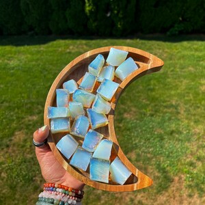 May include: A wooden crescent moon-shaped dish filled with iridescent white opal crystal chips. The dish is held in a hand with several bracelets on the wrist.