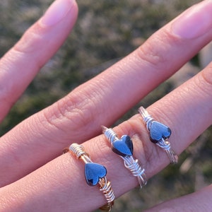 May include: Three gold and silver wire wrapped rings with black heart-shaped stones. The rings are stacked on a finger.