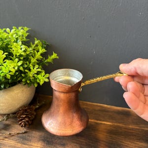 Könnte beinhalten: Eine Kaffeekanne aus Kupfer mit einem breiten, abgerundeten Boden und einem schmalen Hals. Das Innere ist mit einem silberfarbenen Metall ausgekleidet. Sie hat einen langen, strukturierten Messinggriff, der seitlich am Rand befestigt ist.