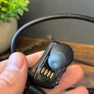 May include: A black Sony Walkman digital music player with a wrap-around neckband design. The device features a prominent R marking for the right side, gold-colored charging contacts, a reset button, and a translucent blue silicone ear tip.