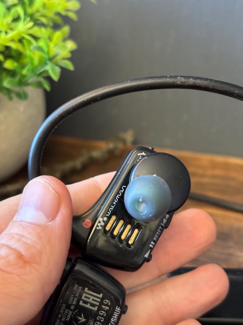 May include: A black Sony Walkman digital music player with a wrap-around neckband design. The device features a prominent R marking for the right side, gold-colored charging contacts, a reset button, and a translucent blue silicone ear tip.