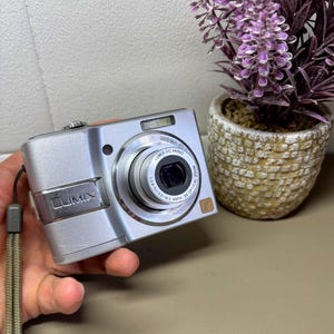 以下が含まれることがあります： 格納式レンズとリストストラップが付いたシルバーのLumixデジタルカメラ。「LUMIX DC VARIO」と「3X OPTICAL ZOOM」の文字がレンズに印刷されています。カメラは手で持たれています。